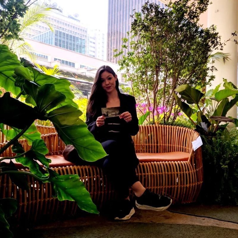 Uma mulher sentada em um sofá de palha, segurando uma foto, cercada por plantas em um ambiente urbano.