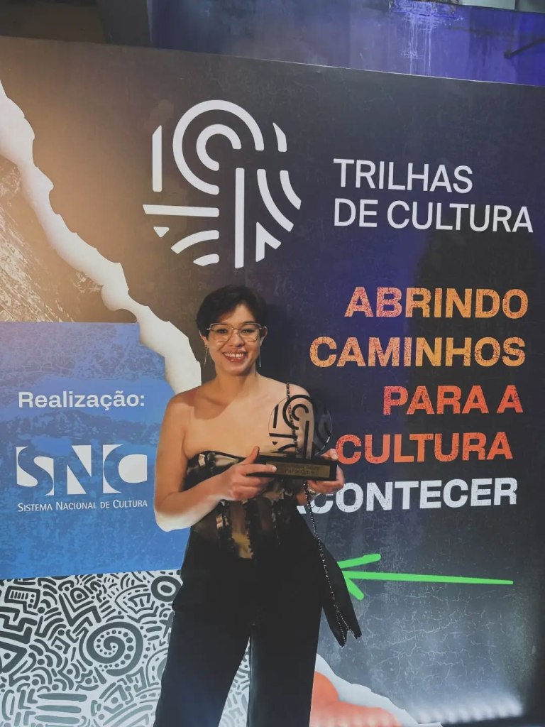 Uma mulher segurando um troféu em frente a um painel com a frase 'Trilhas de Cultura' e 'Abrindo Caminhos para a Cultura'.