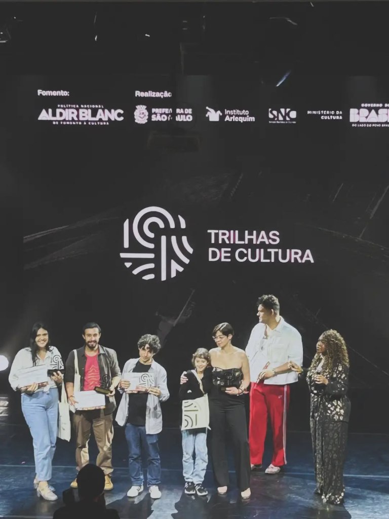 Grupo de pessoas sorrindo em um evento, segurando prêmios e certificados, com um fundo escuro e a frase 'TRILHAS DE CULTURA' em destaque.