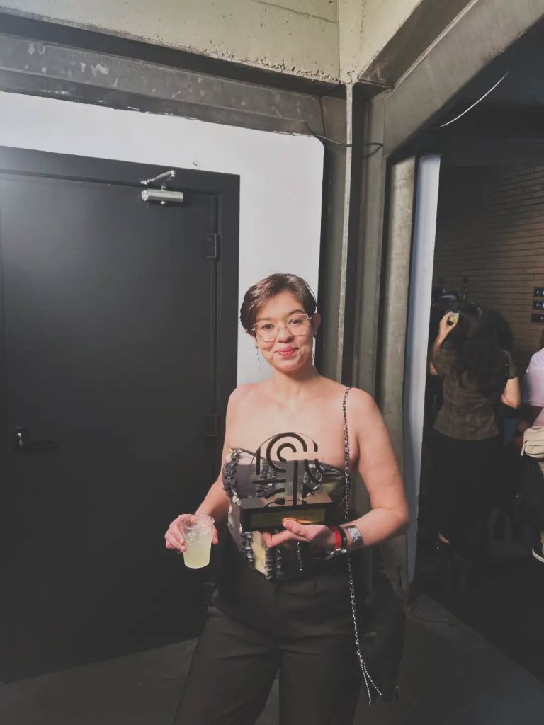 Mulher segurando um troféu e uma bebida em um ambiente interno com uma porta preta ao fundo.