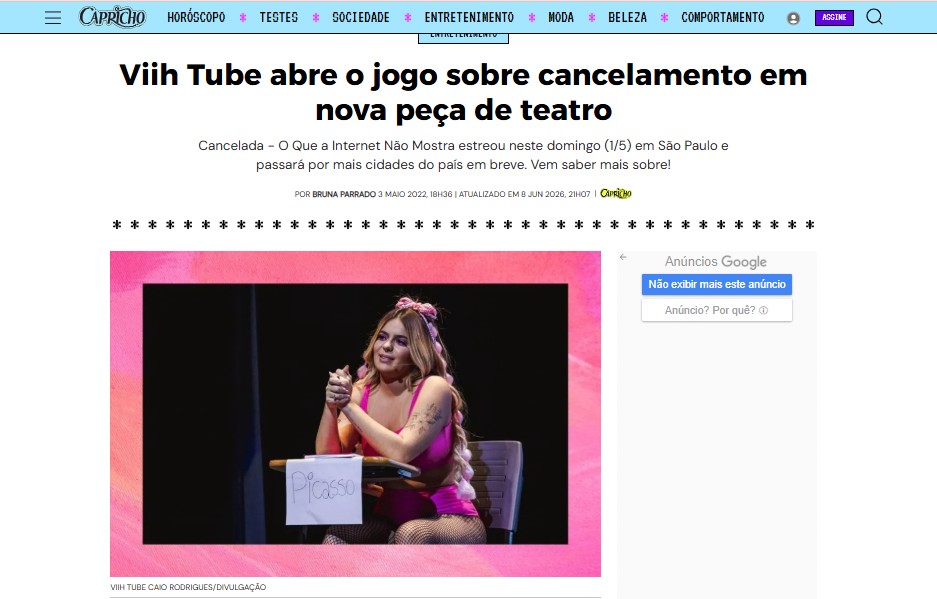 Viihtube em cena de teatro, sentada em uma cadeira com uma placa escrita 'Picasso'. Ela usa um traje colorido e sorri, com luzes rosa ao fundo.